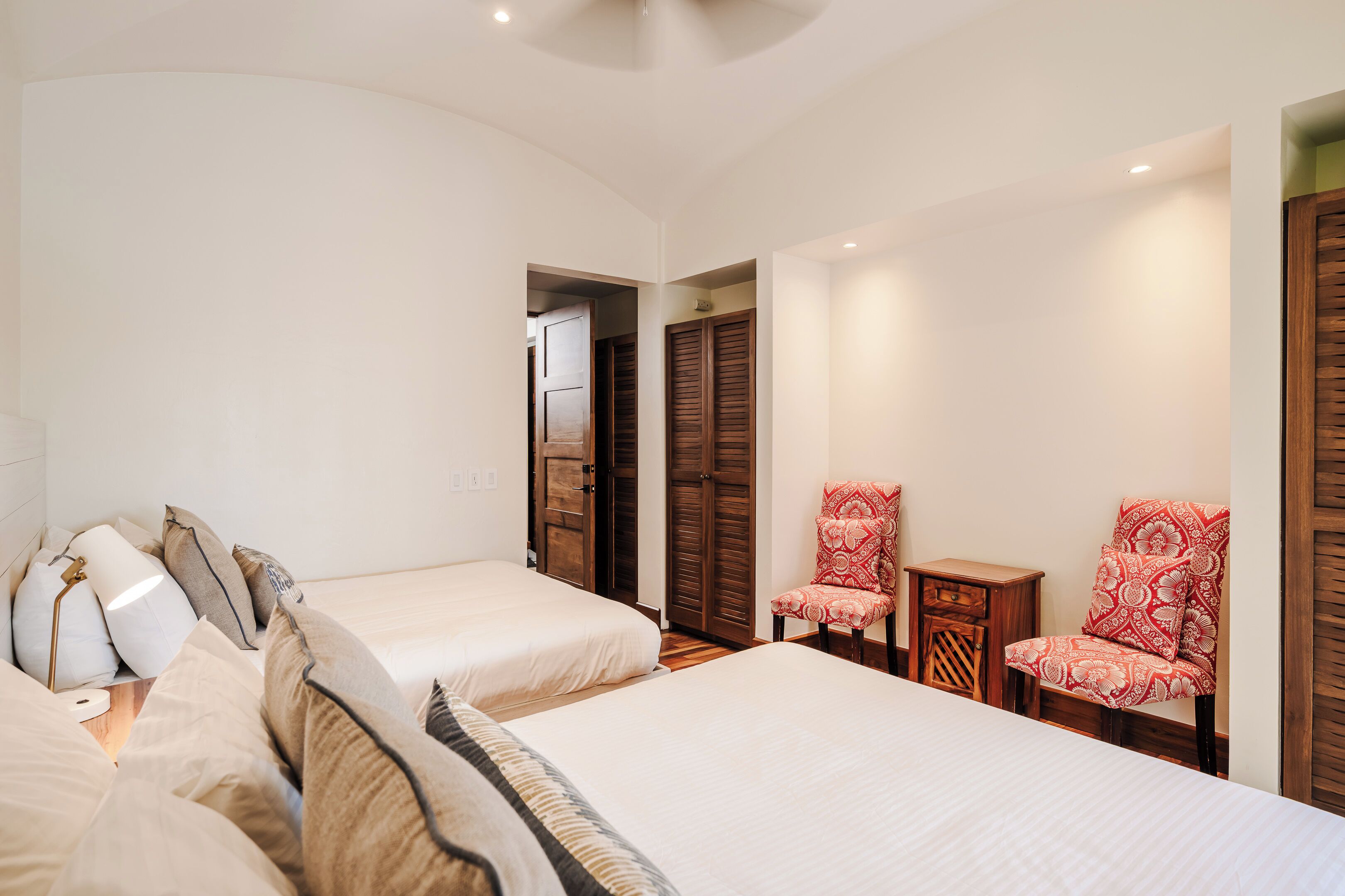 A bedroom with two beds, a wooden wardrobe, a red patterned chair, and a small wooden table.