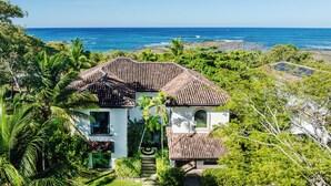 A house with a tiled roof, surrounded by lush greenery and overlooking a beach. - Perfect Family Retreat with the best Sunset views: Luxury Beachfront house w/ Pool & Heated Jacuzzi (Hacienda Pinilla)