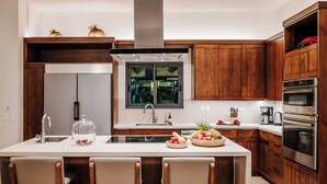 A modern kitchen with wooden cabinets, a stainless steel range, a large island with bar stools, and a window overlooking greenery. - Perfect Family Retreat with the best Sunset views: Luxury Beachfront house w/ Pool & Heated Jacuzzi (Hacienda Pinilla)