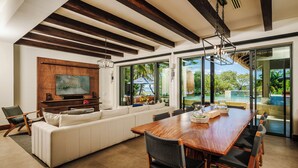 A spacious living area with a wooden dining table, white sofa, and a large TV mounted on the wall. - Perfect Family Retreat with the best Sunset views: Luxury Beachfront house w/ Pool & Heated Jacuzzi (Hacienda Pinilla)