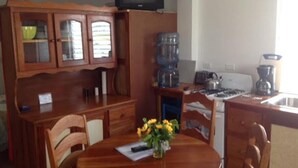 A compact kitchen with wooden cabinets, a dining table with chairs, a water dispenser, and a television above the cabinets. - Trellis House 1 (Caye Caulker)