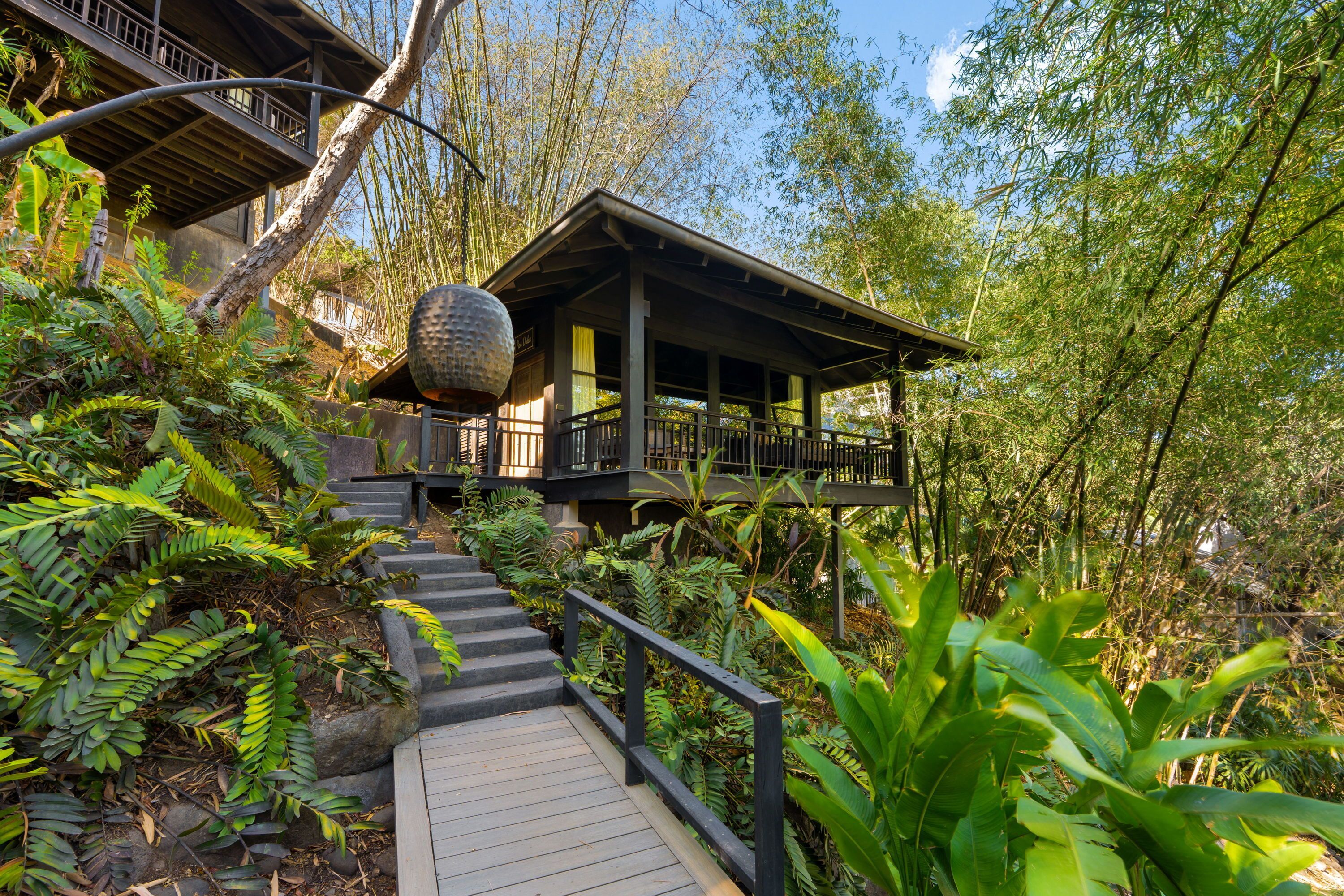 A wooden staircase leading to a covered outdoor area with a railing, surrounded by lush greenery and bamboo trees.