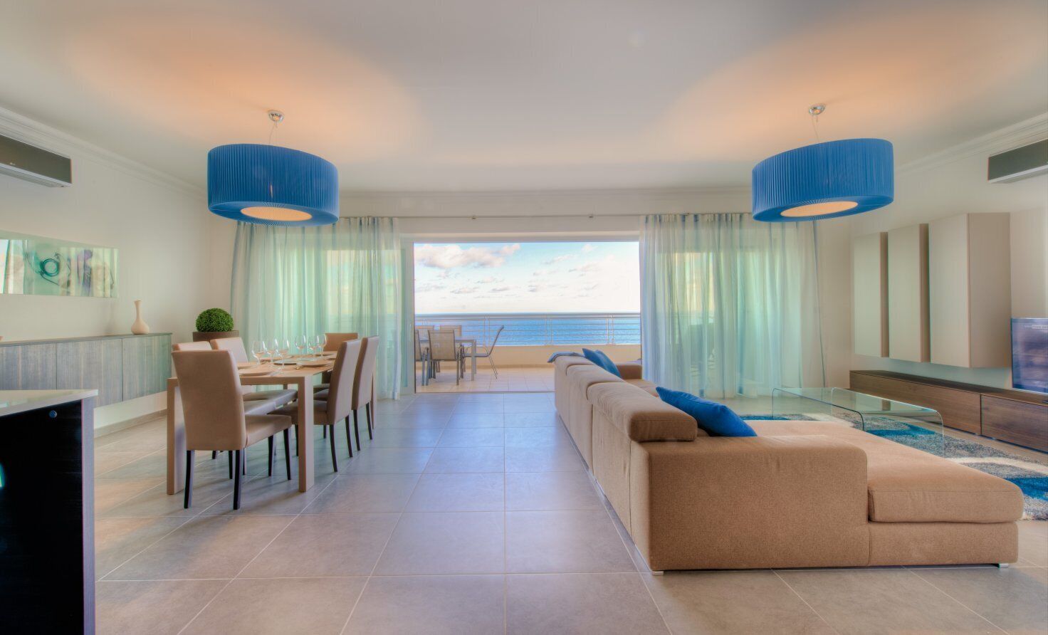A modern living room with a large sofa, dining area, and a view of the sea.
