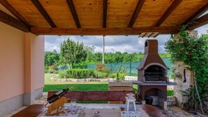 An outdoor wooden table with a bottle, glasses, and a candle, set against a backdrop of a stone patio, a brick oven, and a green garden. - Wonderful private villa with WIFI, private pool, A/C, TV, terrace, pets allowed and panoramic view (Buzet)
