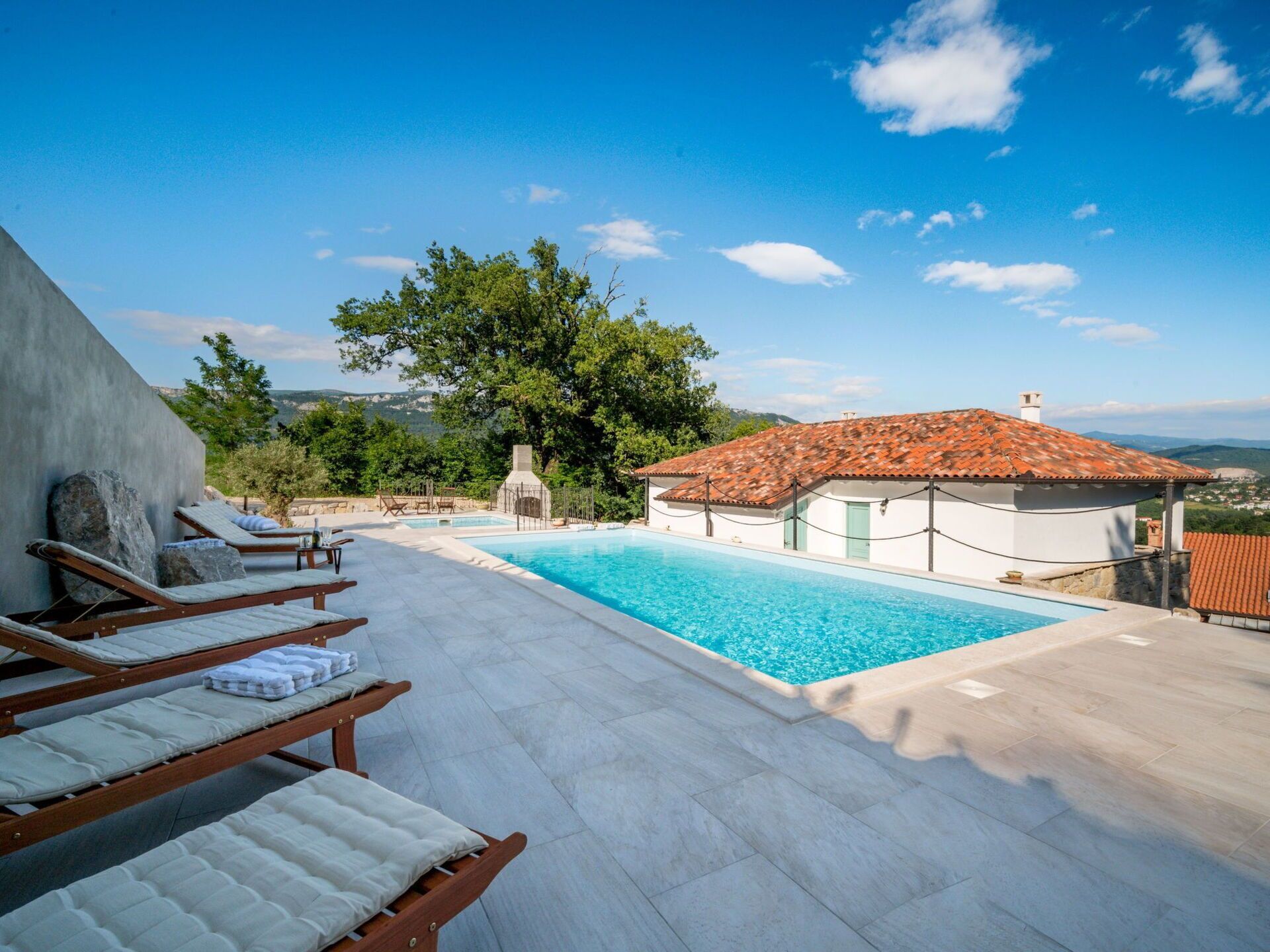 A pool area with lounge chairs, a small building with a tiled roof, and a view of the landscape.