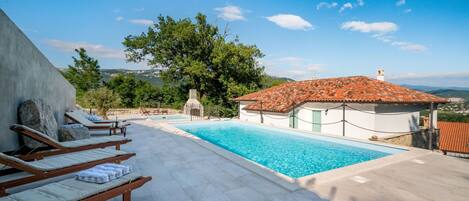 A pool area with lounge chairs, a small building with a tiled roof, and a view of the landscape.
