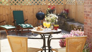 Een terras met rieten stoelen, een ronde tafel met ontbijtartikelen, een barbecue en een houten omheining.