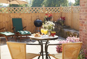 Een terras met rieten stoelen, een ronde tafel met ontbijtartikelen, een barbecue en een houten omheining.