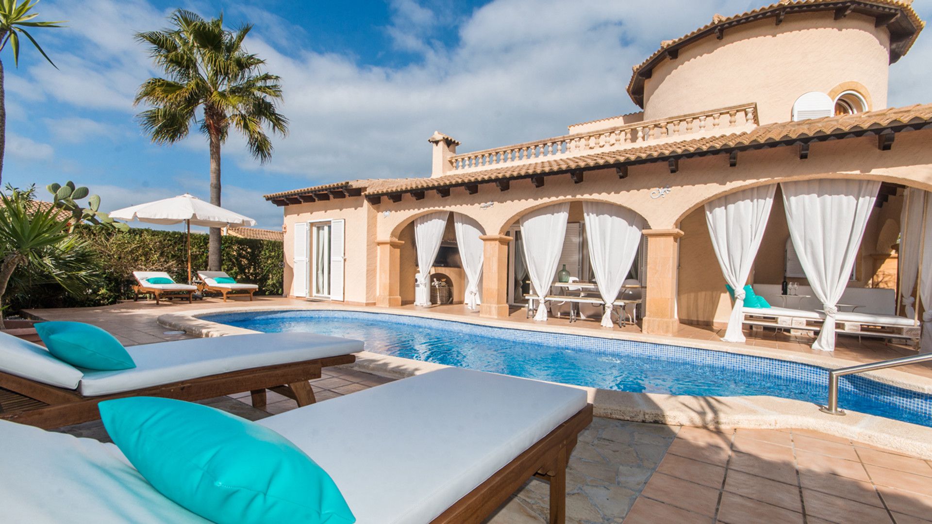 Un área de piscina con sillones de descanso y sombrillas, un edificio con arcos y techo de tejas, y palmeras.