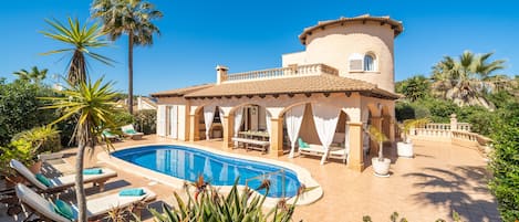 A Mediterranean-style villa with a swimming pool, sun loungers, and lush tropical plants.