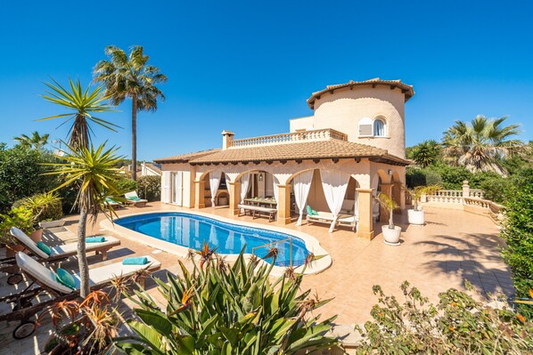 A Mediterranean-style villa with a swimming pool, sun loungers, and lush tropical plants.