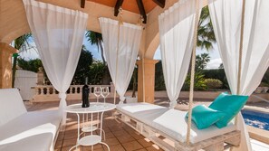 Un área de estar al aire libre cubierta con cortinas blancas, una mesa con una botella y copas, y una piscina al fondo.
