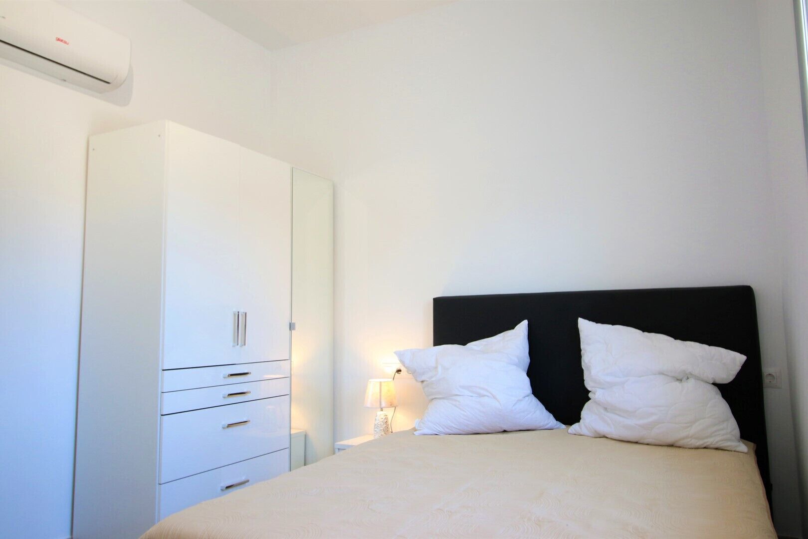 A bedroom with a bed, white wardrobe, bedside lamp, and an air conditioning unit.