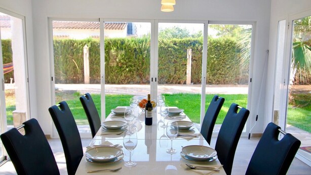 Une table à manger dressée pour six personnes, avec des chaises, de grandes fenêtres et une vue sur un jardin.
