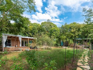 A rustic cabin with a wooden deck and a covered porch, surrounded by lush greenery and a stone pathway.