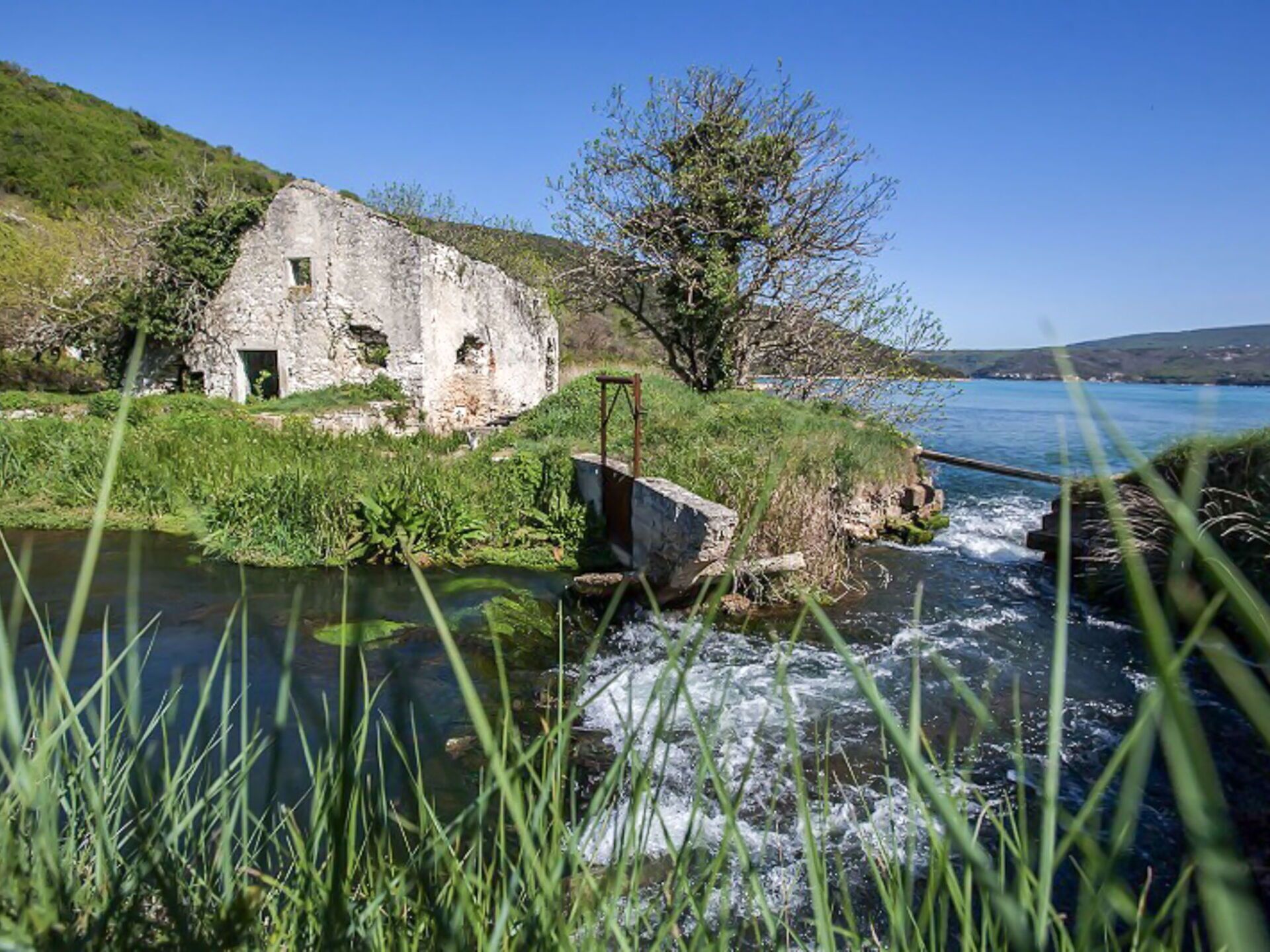 A rustic stone building with a small window, situated by a flowing river with a clear view of the water.