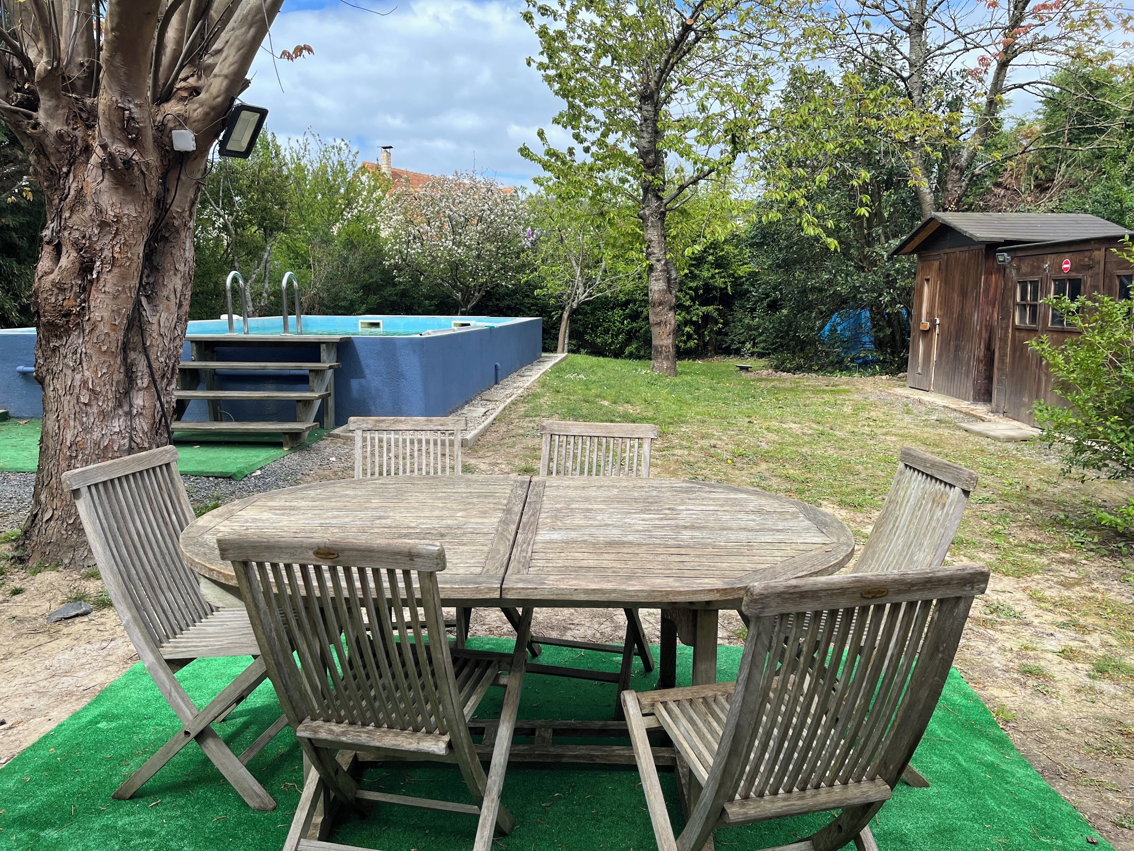 A wooden outdoor dining set with chairs and a table, surrounded by green artificial turf, trees, and a blue structure in the background.