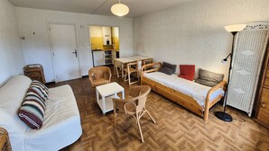 Habitación compacta con sofá, una mesita, una silla, cama y una kitchenette al fondo.