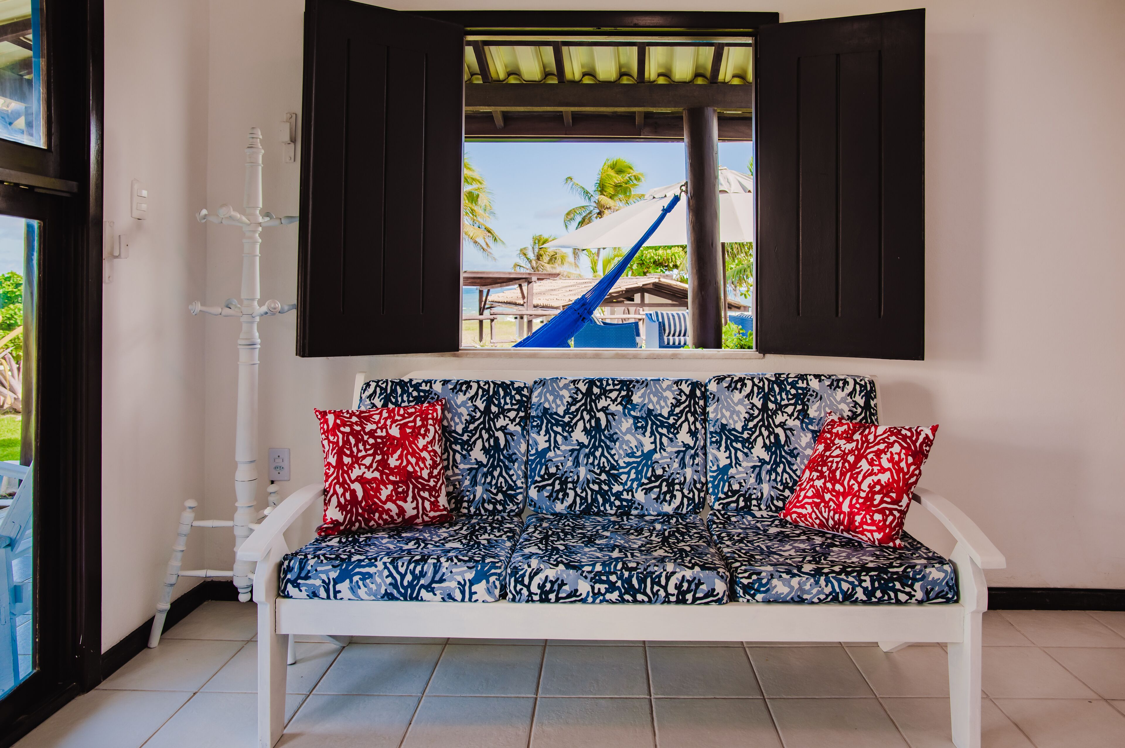 Un sofá blanco con cojines estampados en rojo y azul, una ventana con persianas negras, y vistas a palmeras y un tobogán azul en el exterior.