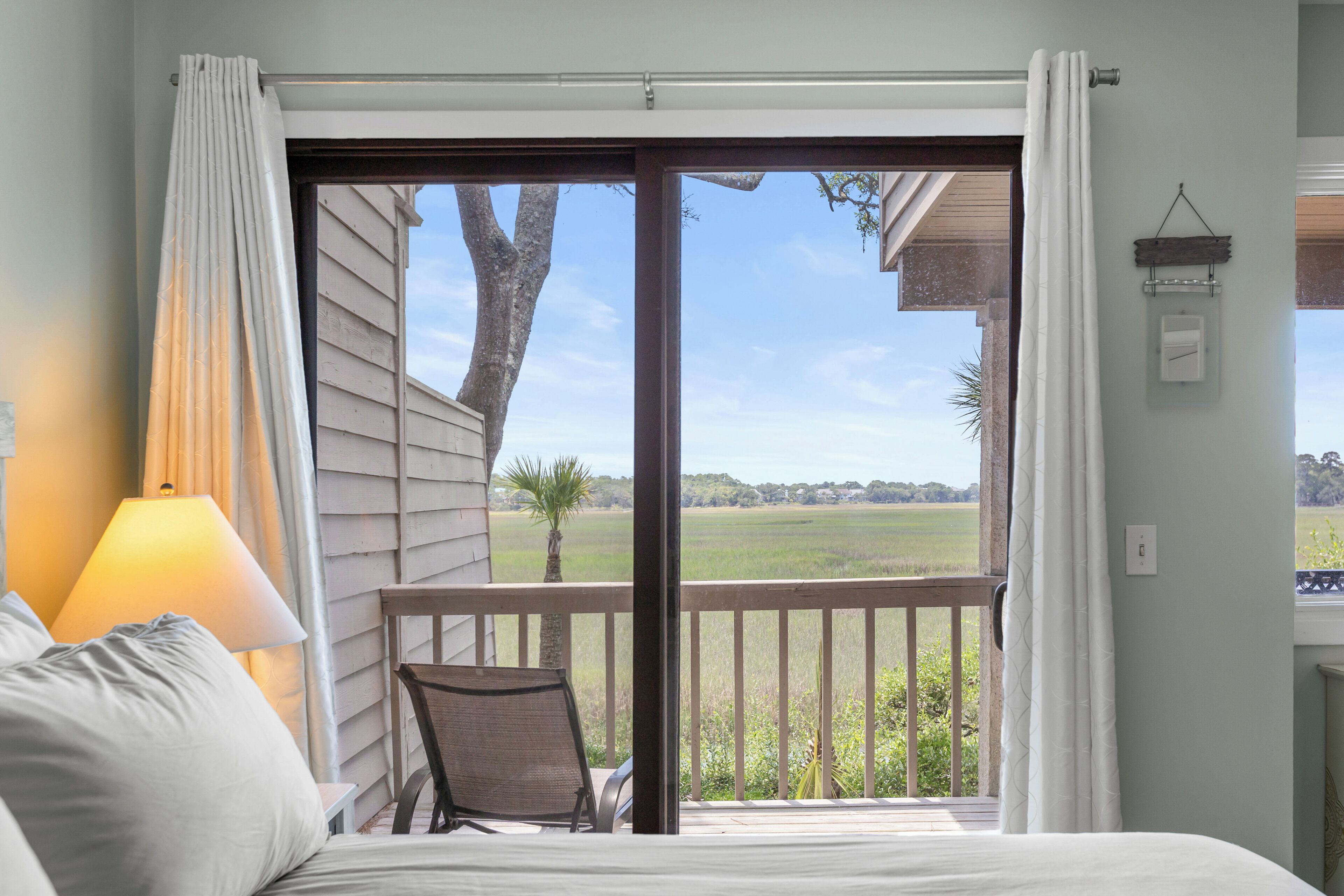 Un dormitorio con cama, una silla, una lámpara y vistas a un paisaje verde a través de un ventanal corredero.
