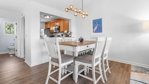 A dining area with a wooden table and white chairs, a kitchen with wooden cabinets, and a hanging light fixture.
