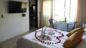 Une chambre d’hôtel avec un lit, un téléviseur fixé au mur, une fenêtre avec des rideaux et une petite table sur laquelle se trouvent une bouteille et des fleurs.