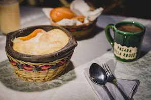 A table with a basket containing bread, a mug labeled "PERSADA JILLA ROUSE," and a spoon and fork on a tablecloth.