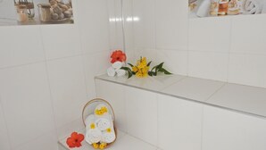 Une salle de bain aux murs recouverts de carreaux blancs, une étagère en verre où sont posés des articles de toilette, et un panier rempli de serviettes roulées, agrémenté de fleurs.