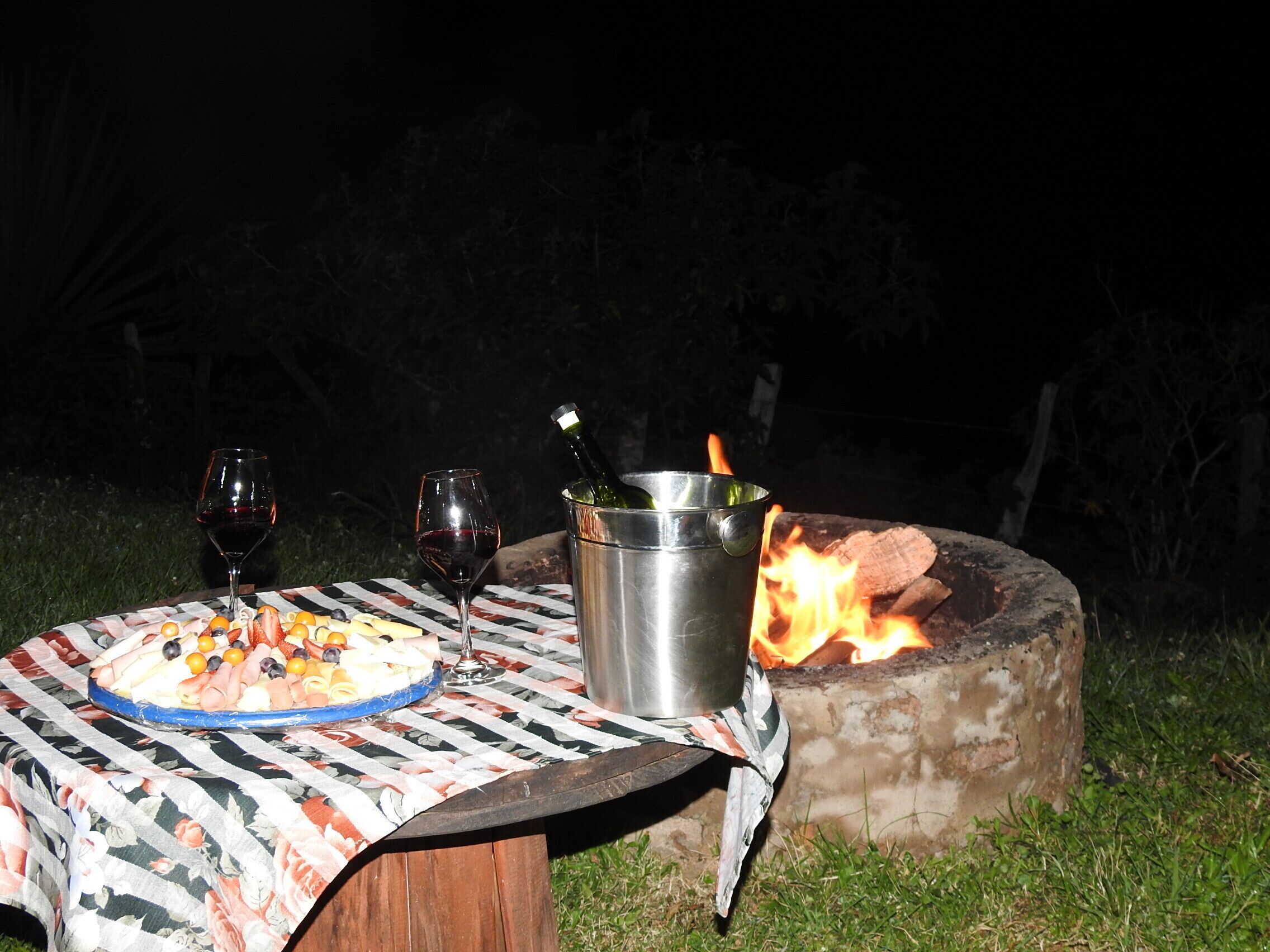 An outdoor dining setup with a table, two wine glasses, a metal bucket, and a campfire.