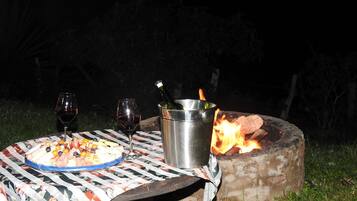 An outdoor dining setup with a table, two wine glasses, a metal bucket, and a campfire.