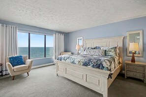 A bedroom with a large bed, a chair, a nightstand, a lamp, and a window with a view of the ocean.