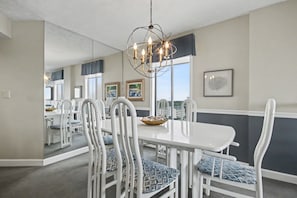 Un coin repas avec une table et des chaises blanches, un lustre et des œuvres d’art encadrées accrochées aux murs.