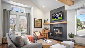 A modern living room with a fireplace, a television mounted above it, and a large window providing a view of the outdoors. - The Moose Lodge - New 2BR, Walk to PCMR / Main St! (Park City)