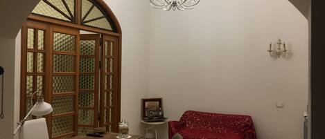 A room with a wooden desk, a chair, a red sofa, a chandelier, and a large arched doorway.