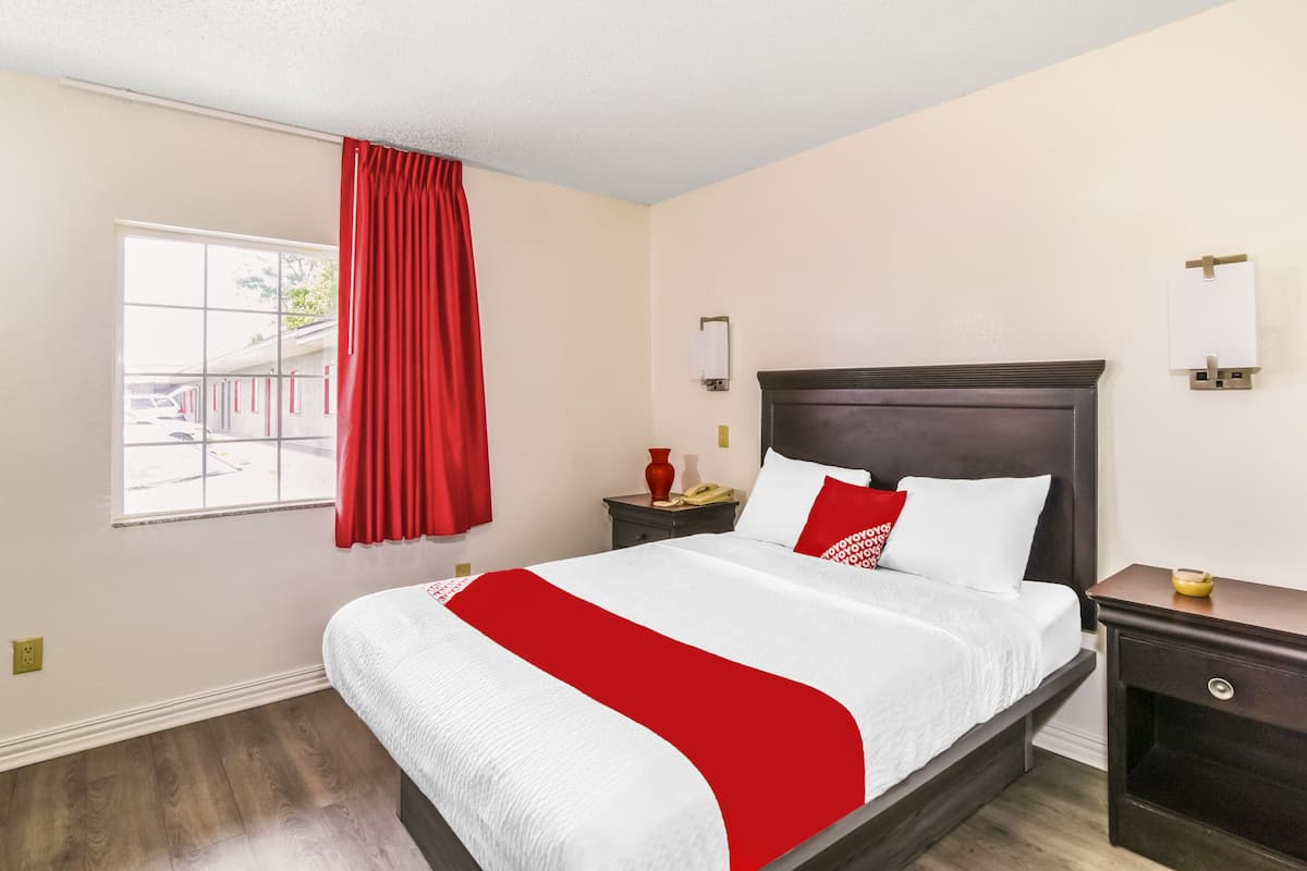 A hotel room with a bed, a nightstand, a window with red curtains, and a wall-mounted light fixture.