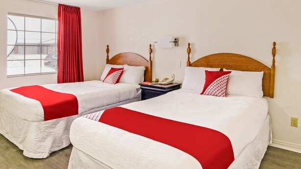 Una habitación de hotel con dos camas, cada una con una colcha roja y blanca, un cabecero de madera, una mesita de noche con un teléfono y una ventana con cortinas rojas.