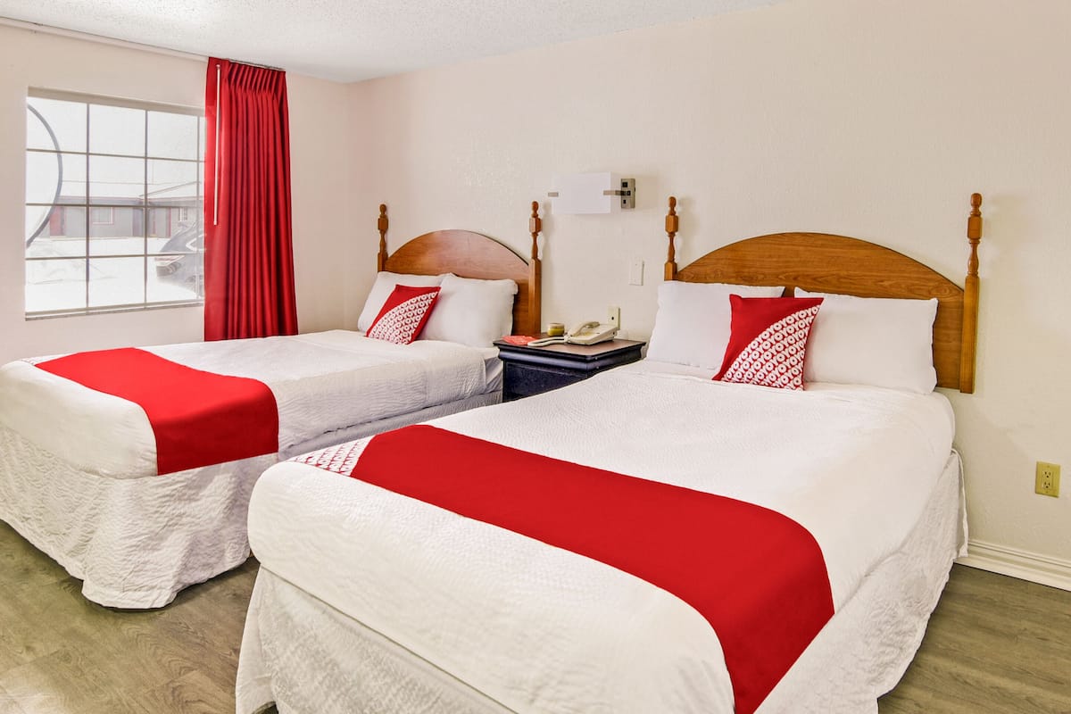 A hotel room with two beds, each with a red and white bedspread, a wooden headboard, a nightstand with a telephone, and a window with red curtains.