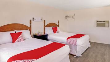 A hotel room with two beds, each with a wooden headboard, white bedding, and red accents. There is a bedside table with a lamp, a telephone, and a small decorative item. The room has a light-colored wall, a ceiling light, and a wall-mounted air conditioning unit.
