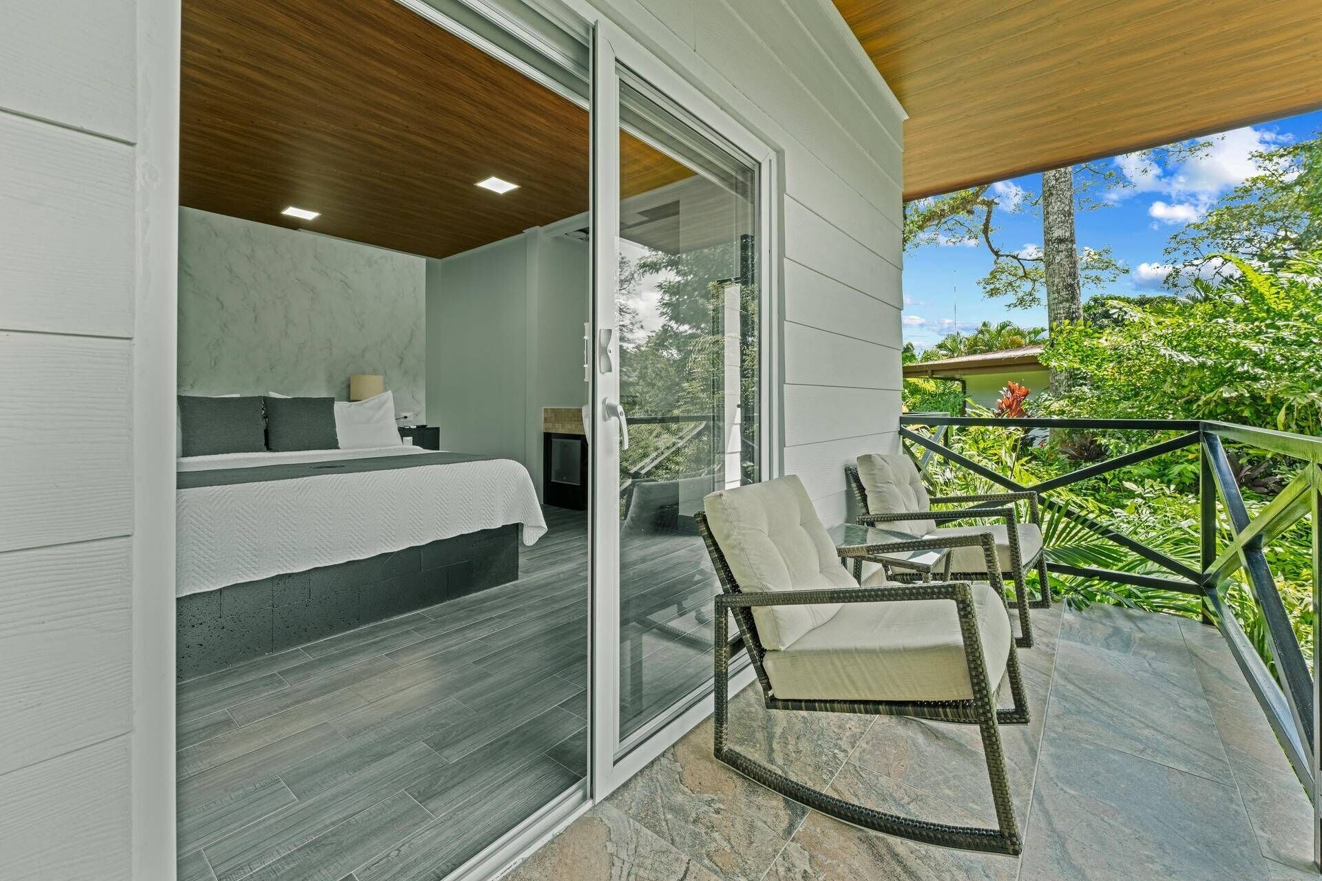 A modern bedroom with a large bed, a nightstand, and a glass door leading to a balcony with wicker chairs and a view of greenery.