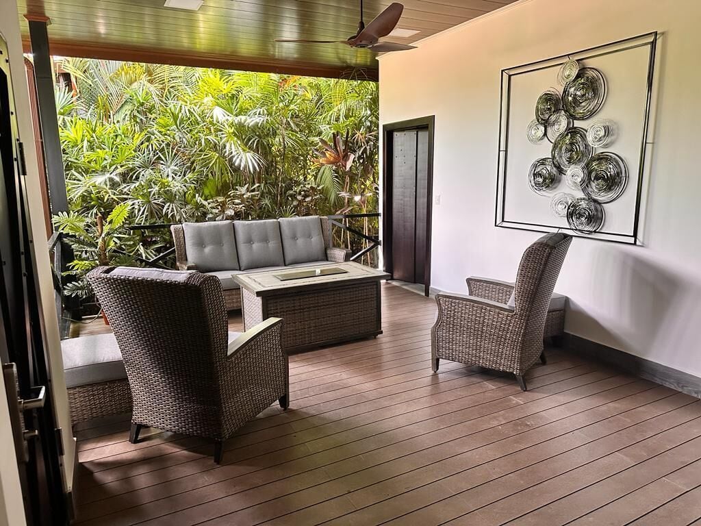 A balcony with wicker furniture, a glass-top table, and a wall-mounted abstract art piece.