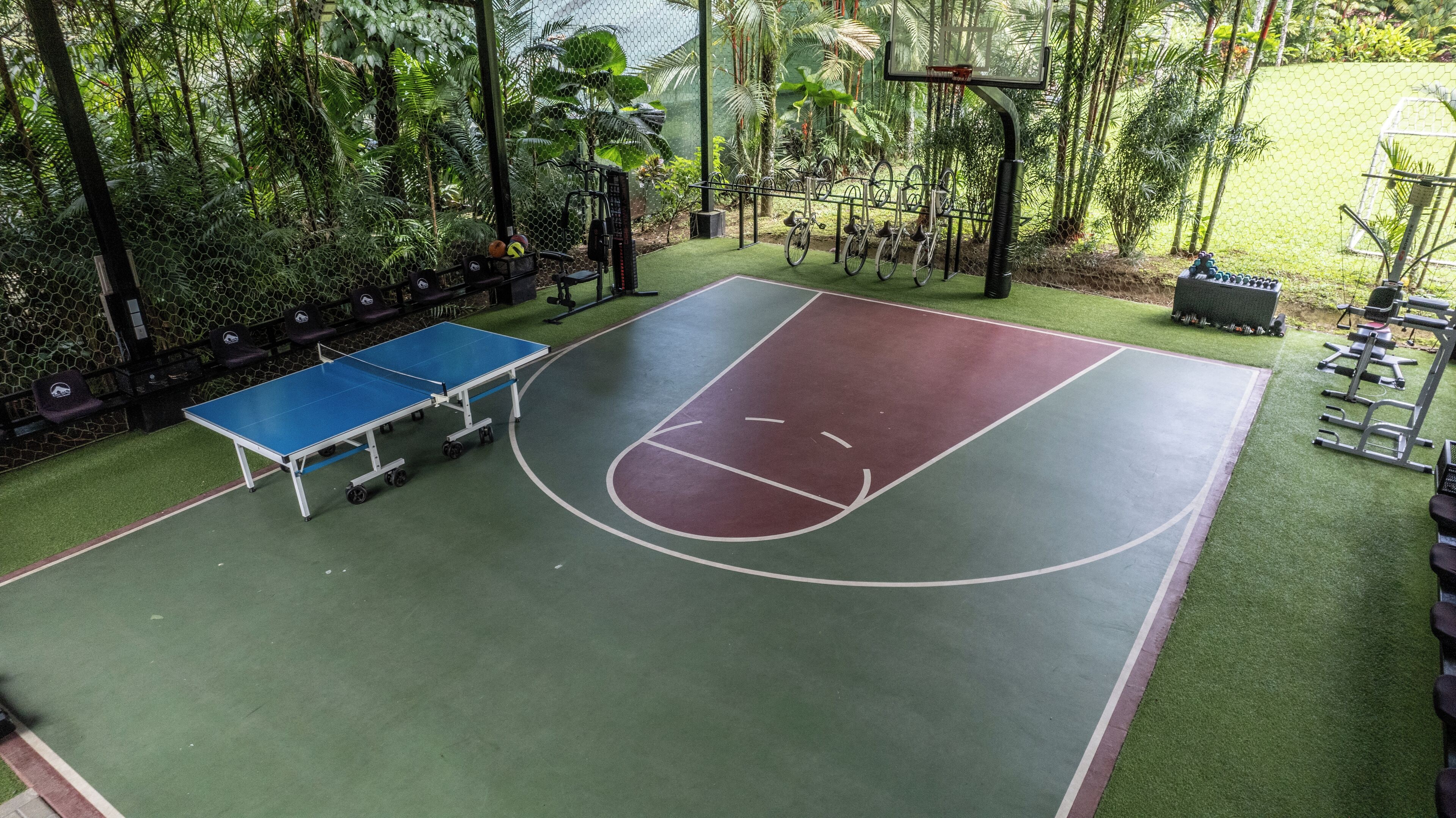 An outdoor basketball court with a blue table tennis table, surrounded by greenery and exercise equipment.