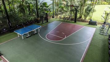 An outdoor basketball court with a blue table tennis table, surrounded by greenery and exercise equipment.