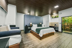 Une chambre d’hôtel moderne avec un plafond en bois, un grand lit recouvert d’une couverture verte, un bureau avec un ordinateur et une vue sur la verdure à travers une porte vitrée.