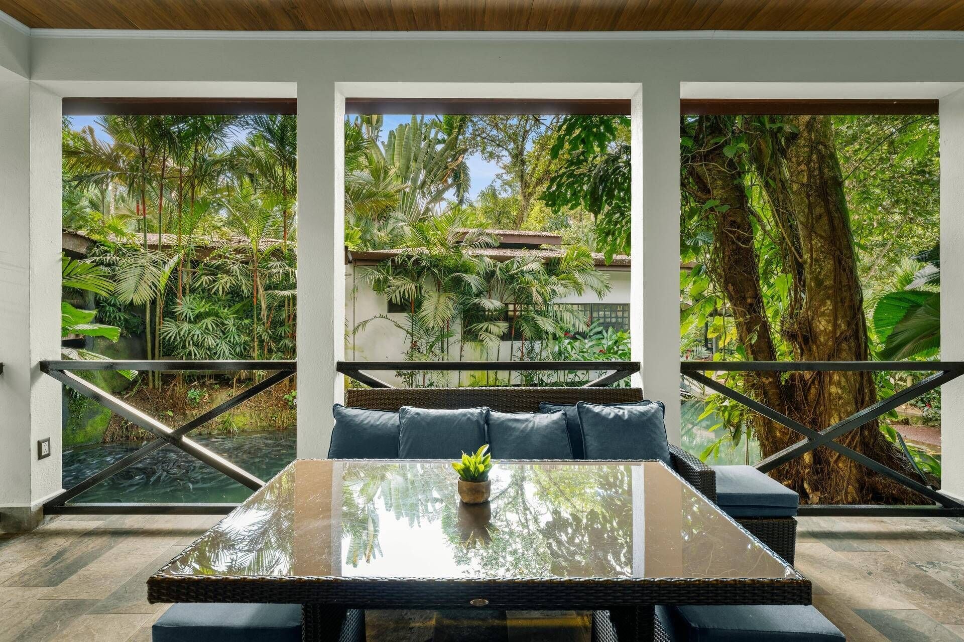 Un espace de détente extérieur moderne, avec une table en verre et des coussins, entouré d’une végétation tropicale luxuriante et offrant une vue sur une maison traditionnelle.