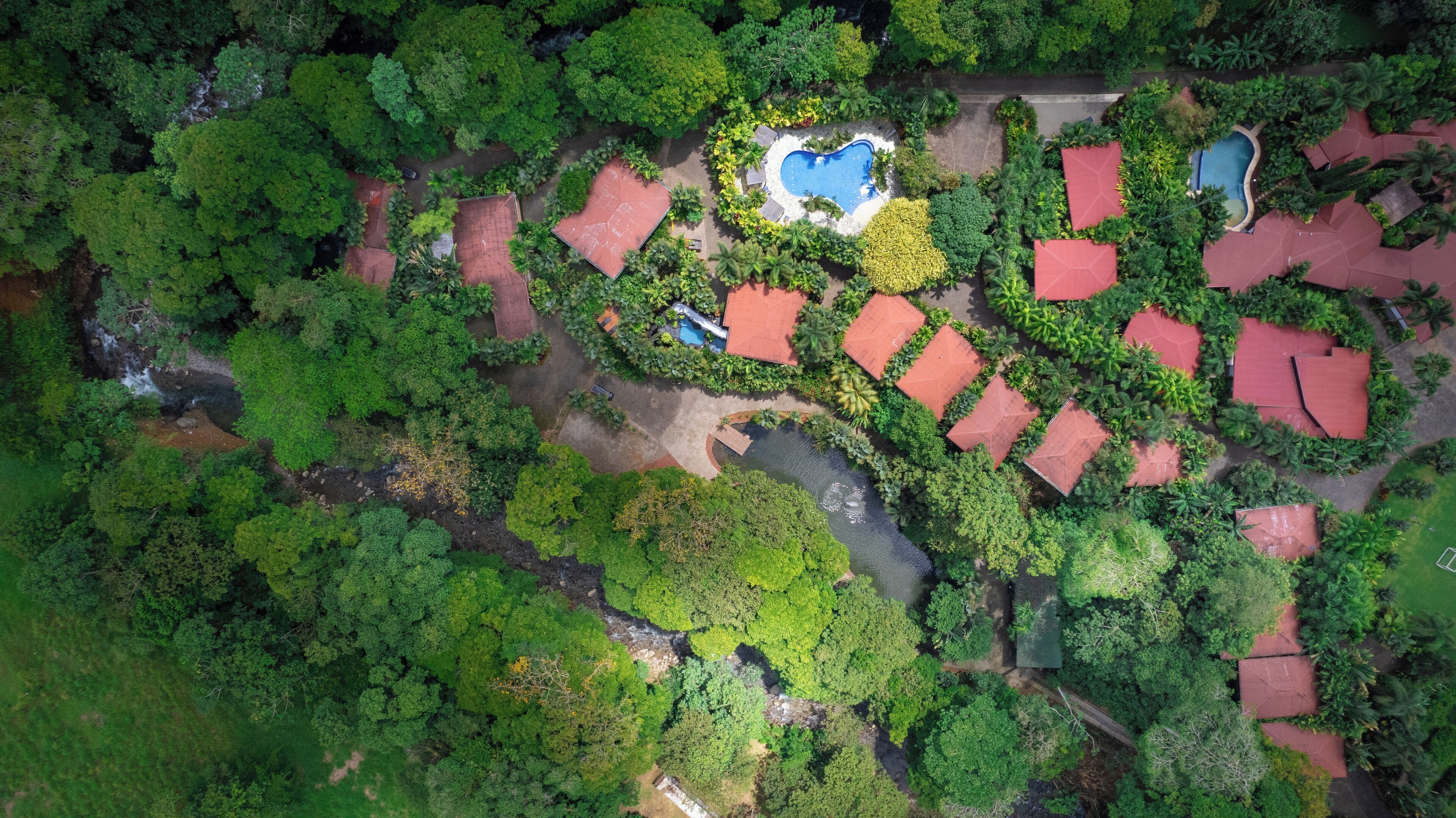 An aerial view of a resort with multiple red-roofed buildings, a central swimming pool, and lush greenery surrounding the area.