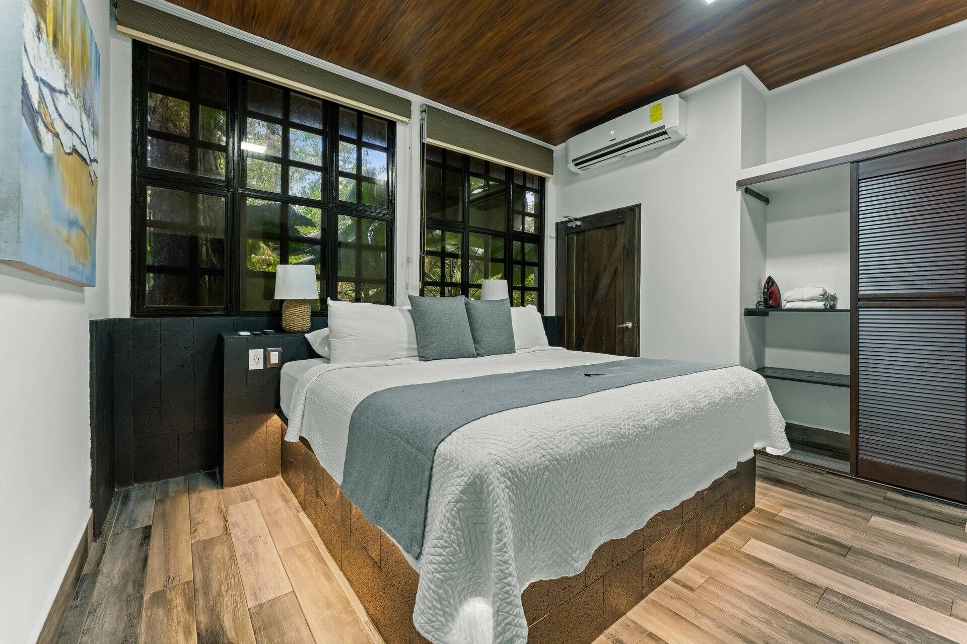 A bedroom with a bed, nightstands, a lamp, a window with a view, an air conditioning unit, and a sliding door closet.