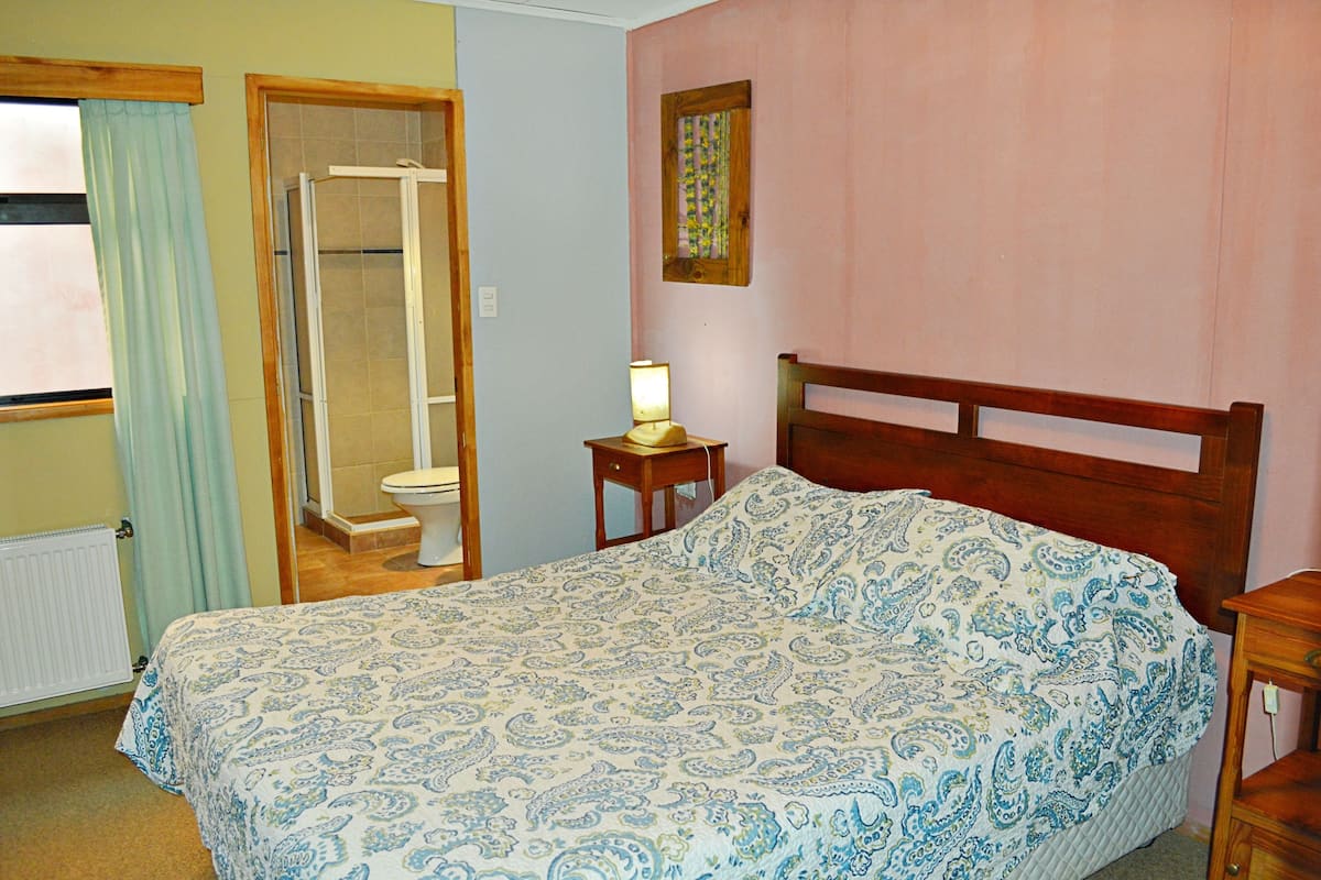 A bedroom with a bed, a wooden headboard, a bedside table with a lamp, a window with curtains, and a bathroom with a shower and toilet.