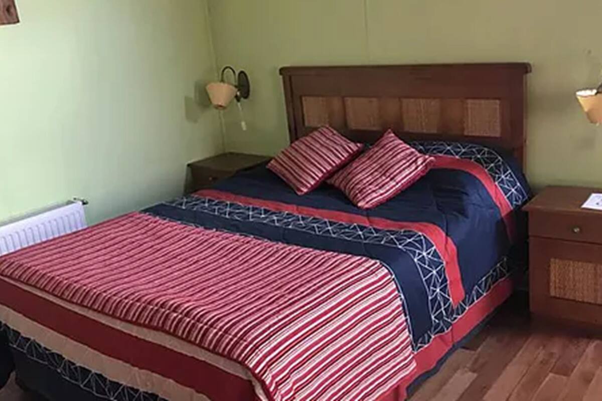 A bed with a patterned bedspread, a nightstand with a lamp, and a wall-mounted telephone.