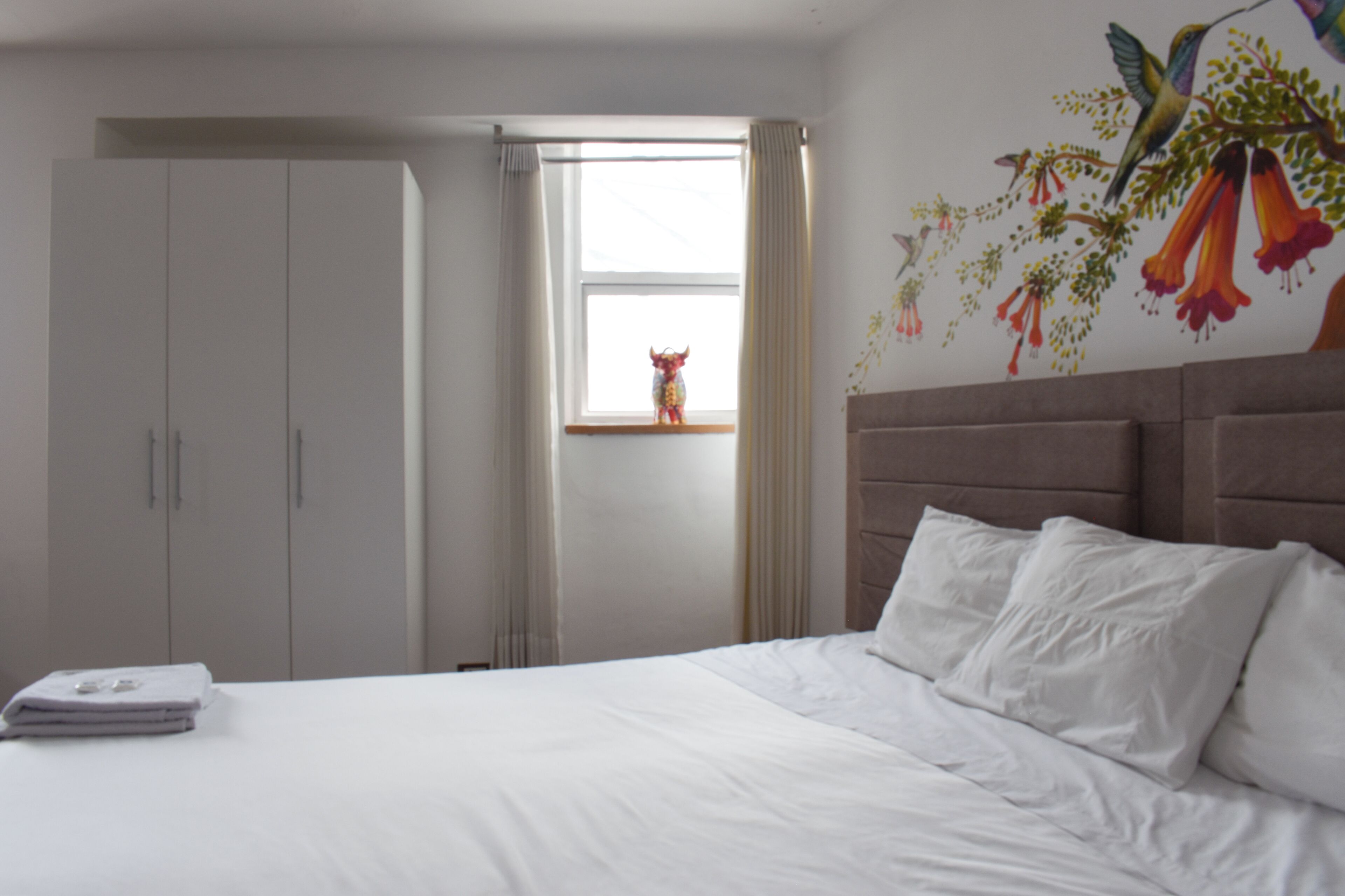 Habitación de hotel con cama, armario, ventana con cortinas y un mural en la pared.