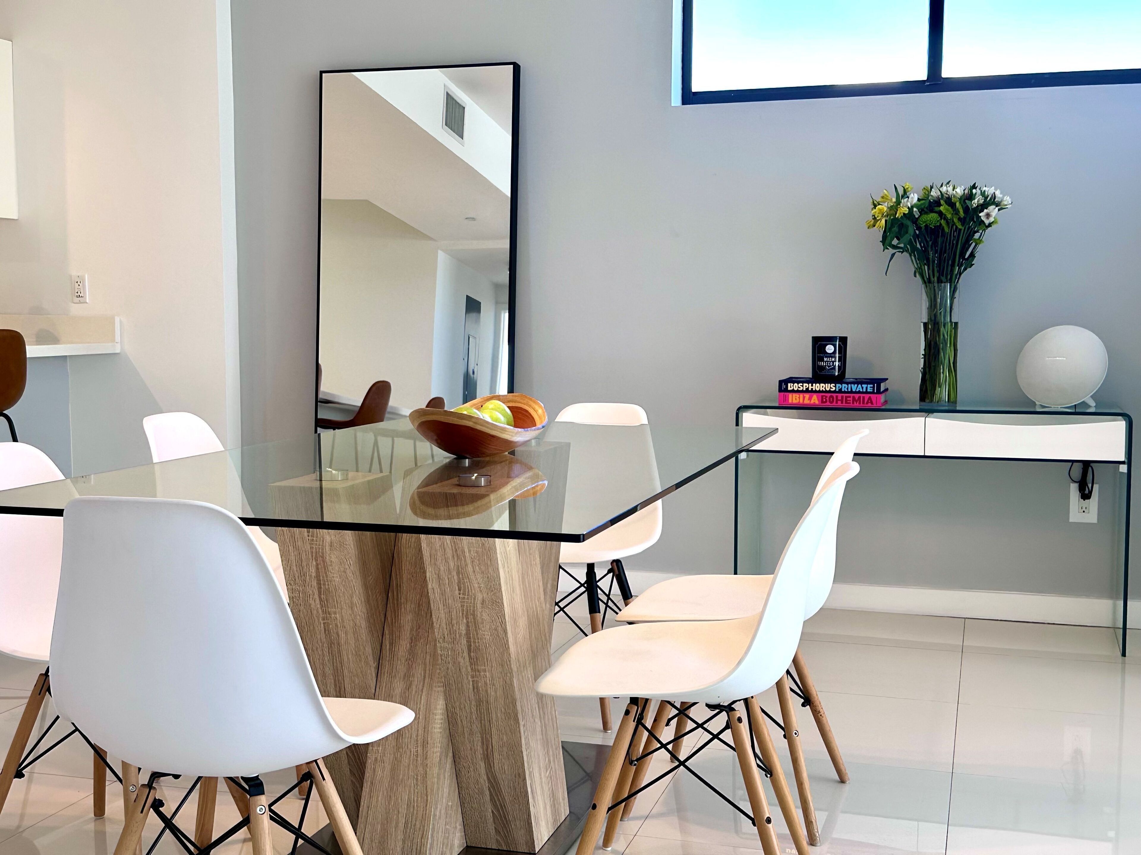 A modern dining area with a glass table, white chairs, a wooden bowl with fruit, a mirror, and a vase with flowers on a console table.